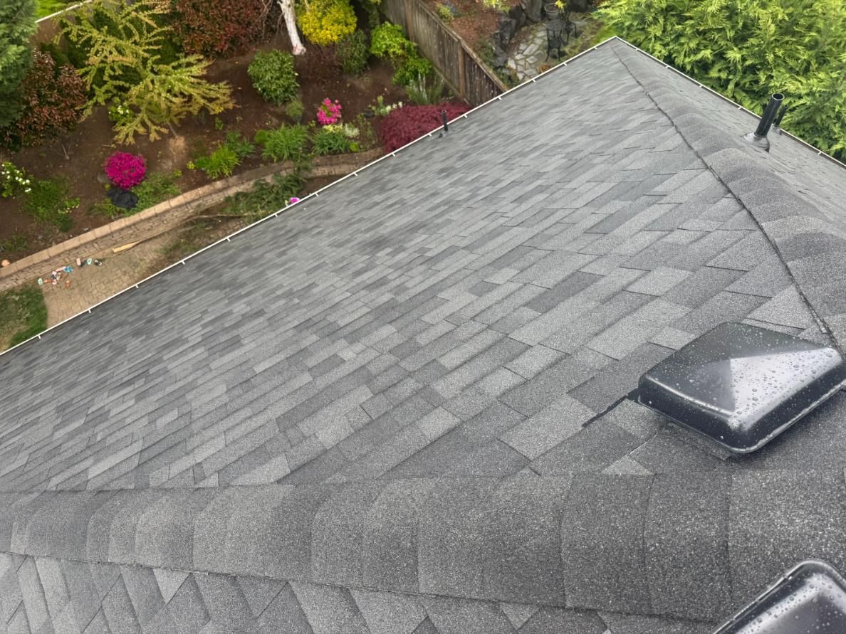 Dark gray asphalt shingle roof, view from above, with garden in the background.