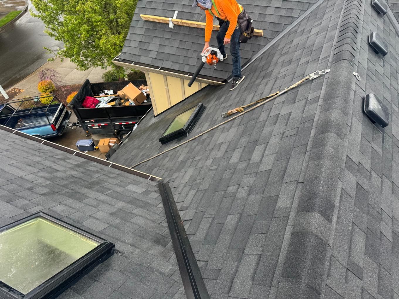 Roofer using a chainsaw on a dark shingled roof, near a skylight, with a construction dumpster below.