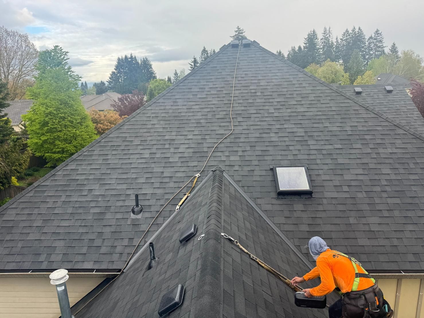 Person on a roof repairs a dark shingle roof. Setting is outdoors with trees under a cloudy sky.