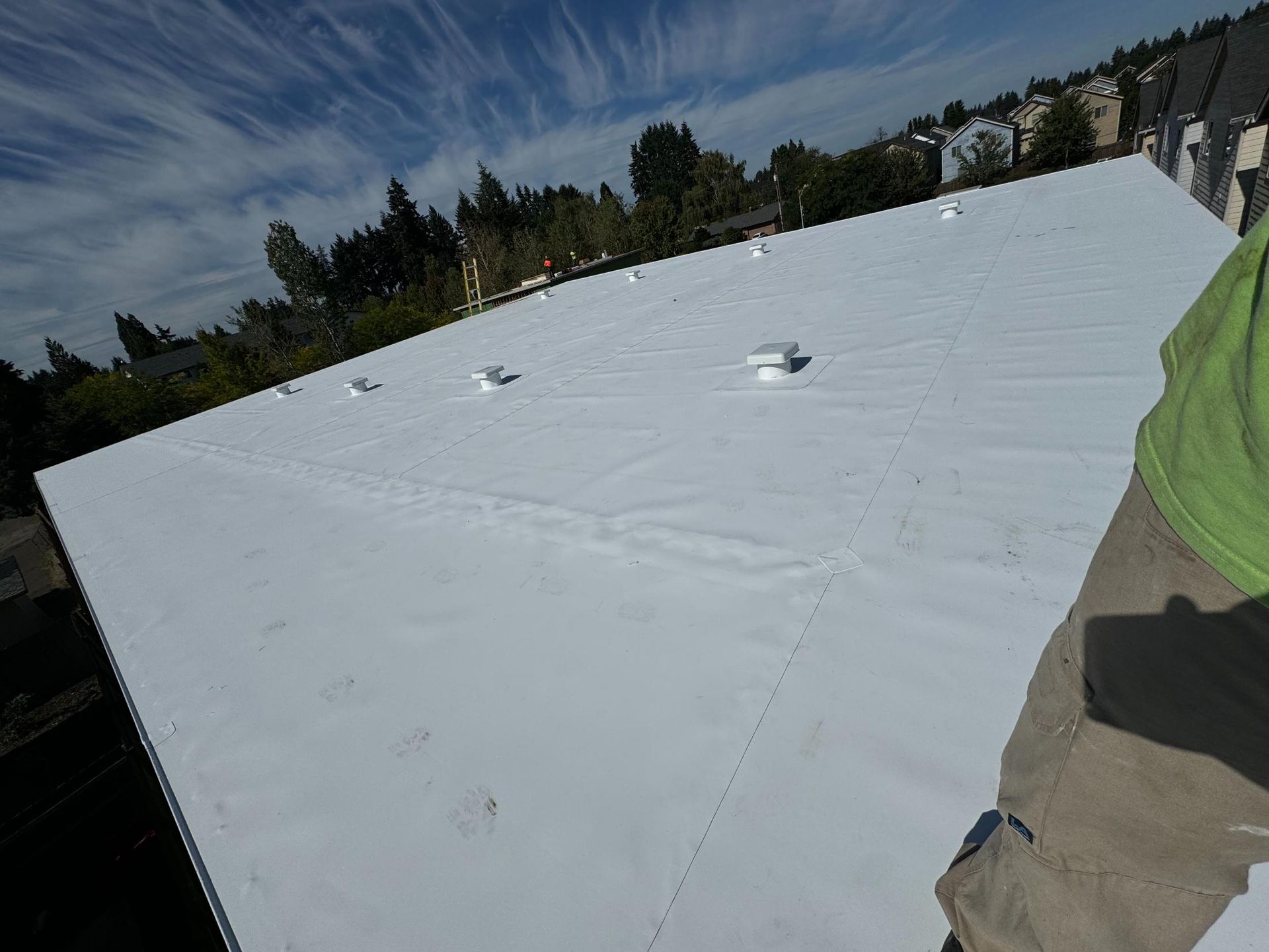 White flat roof with vents and a person in a green shirt and tan pants standing on it under a blue sky.