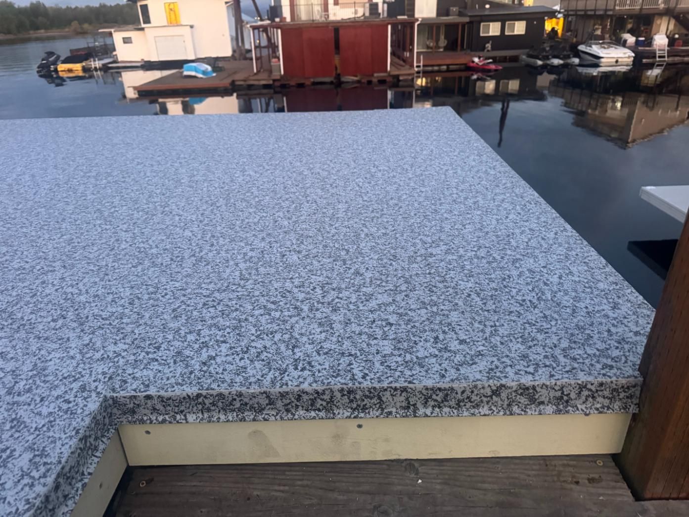 Close-up of a granite-covered platform with water and houseboats in the background. The platform is gray and white.