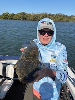 Person in hooded shirt holds a large, flat fish on a boat, near water and trees.