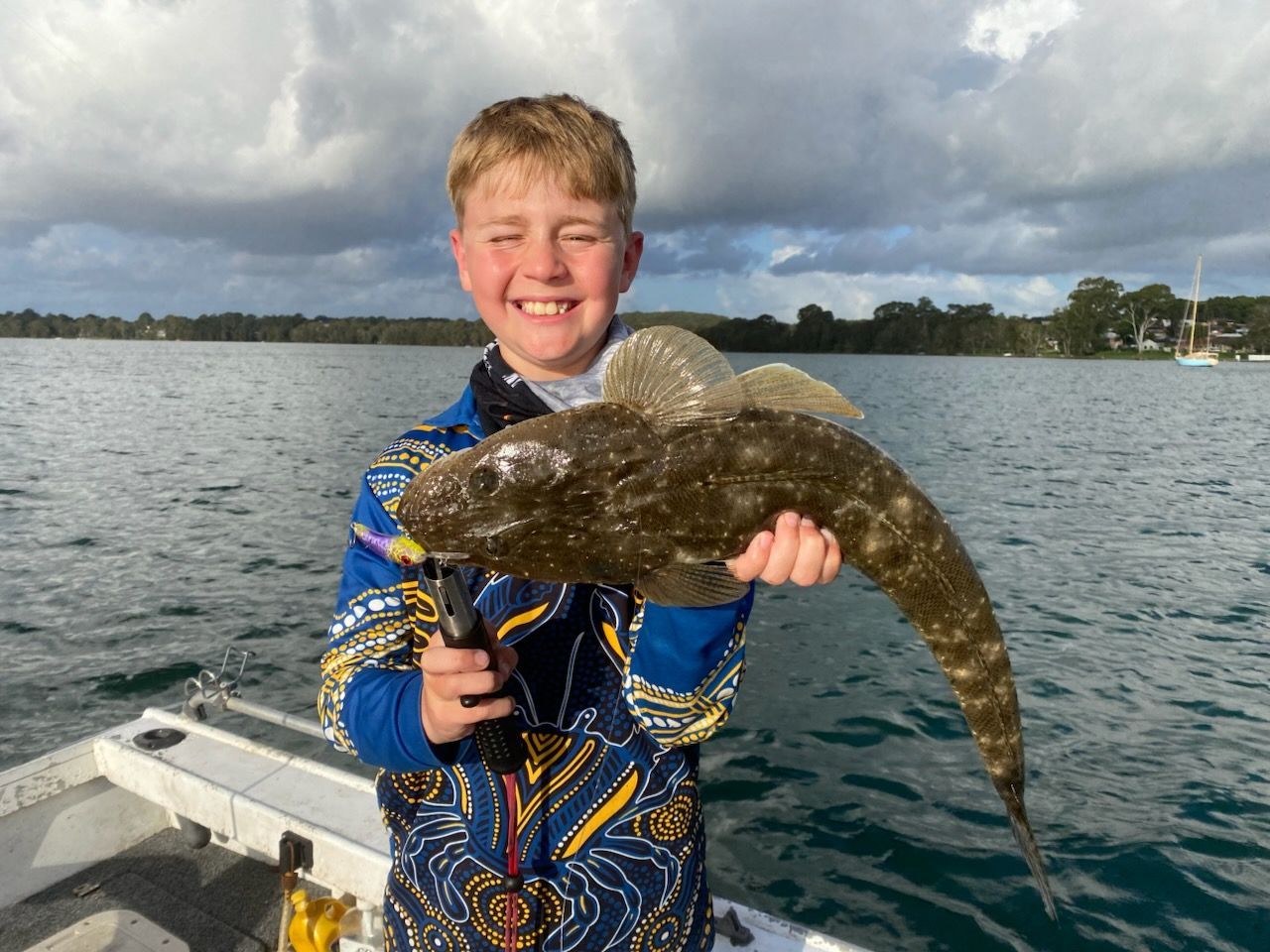 Boy smiling on a boat holding a large, speckled fish. Overcast sky and water in the background.