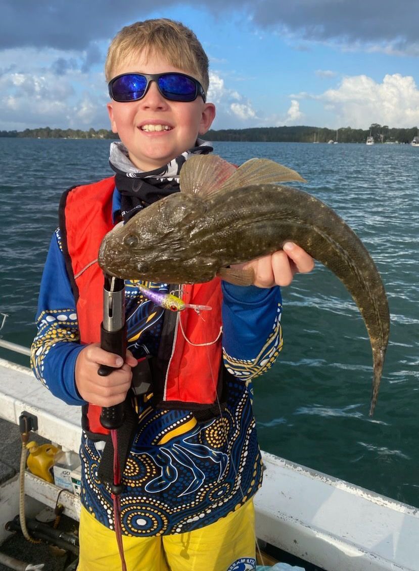 Boy on a boat smiles, holding a flathead fish. Wearing sunglasses, life vest, and patterned clothing. Outdoors, water.