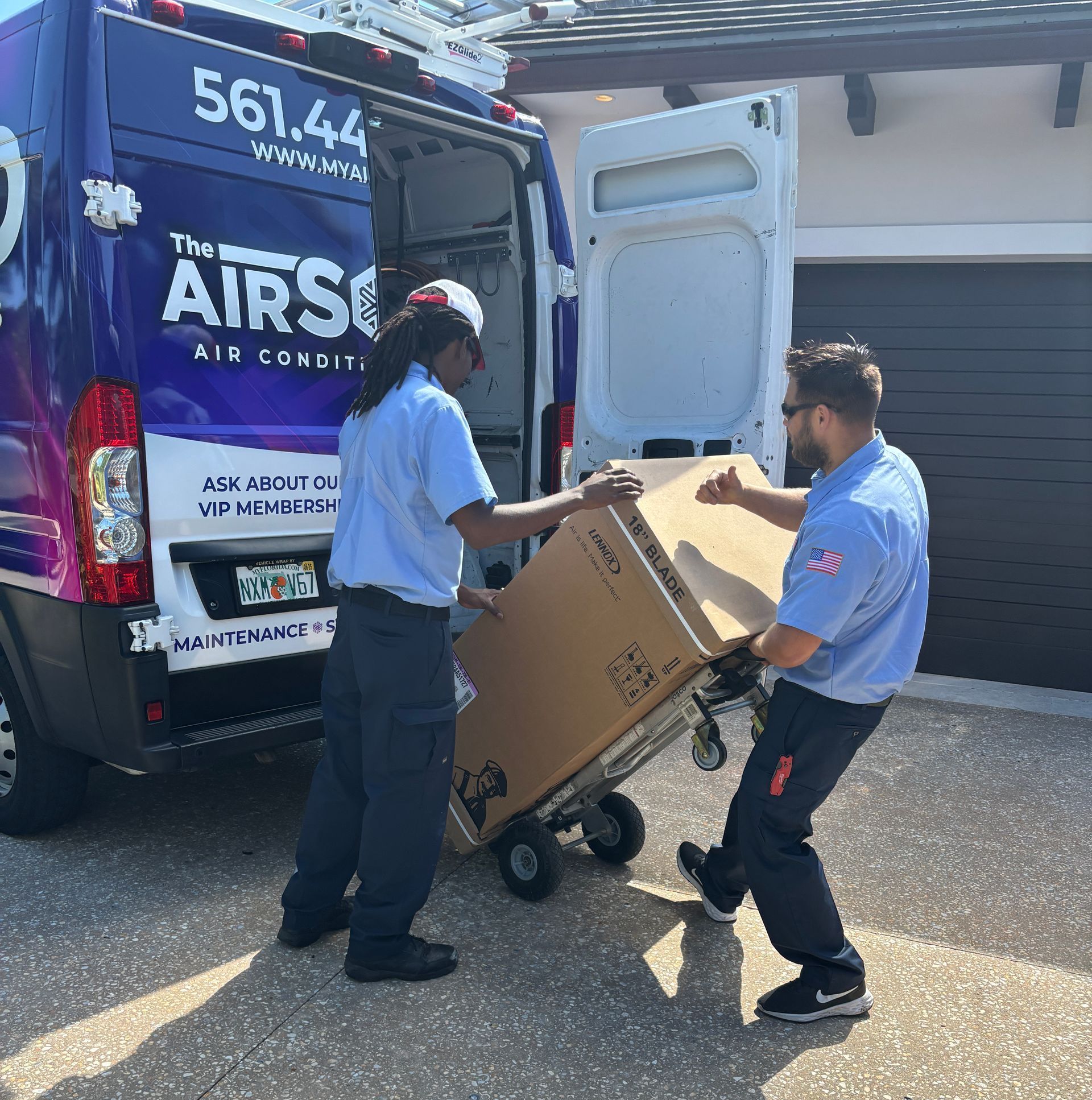 Two men are pushing a large box on a cart in front of an air conditioning van.