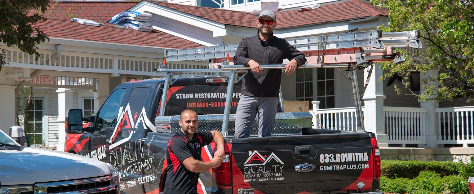 Two men in a truck with ladders, in front of a house. One man leans, the other stands.