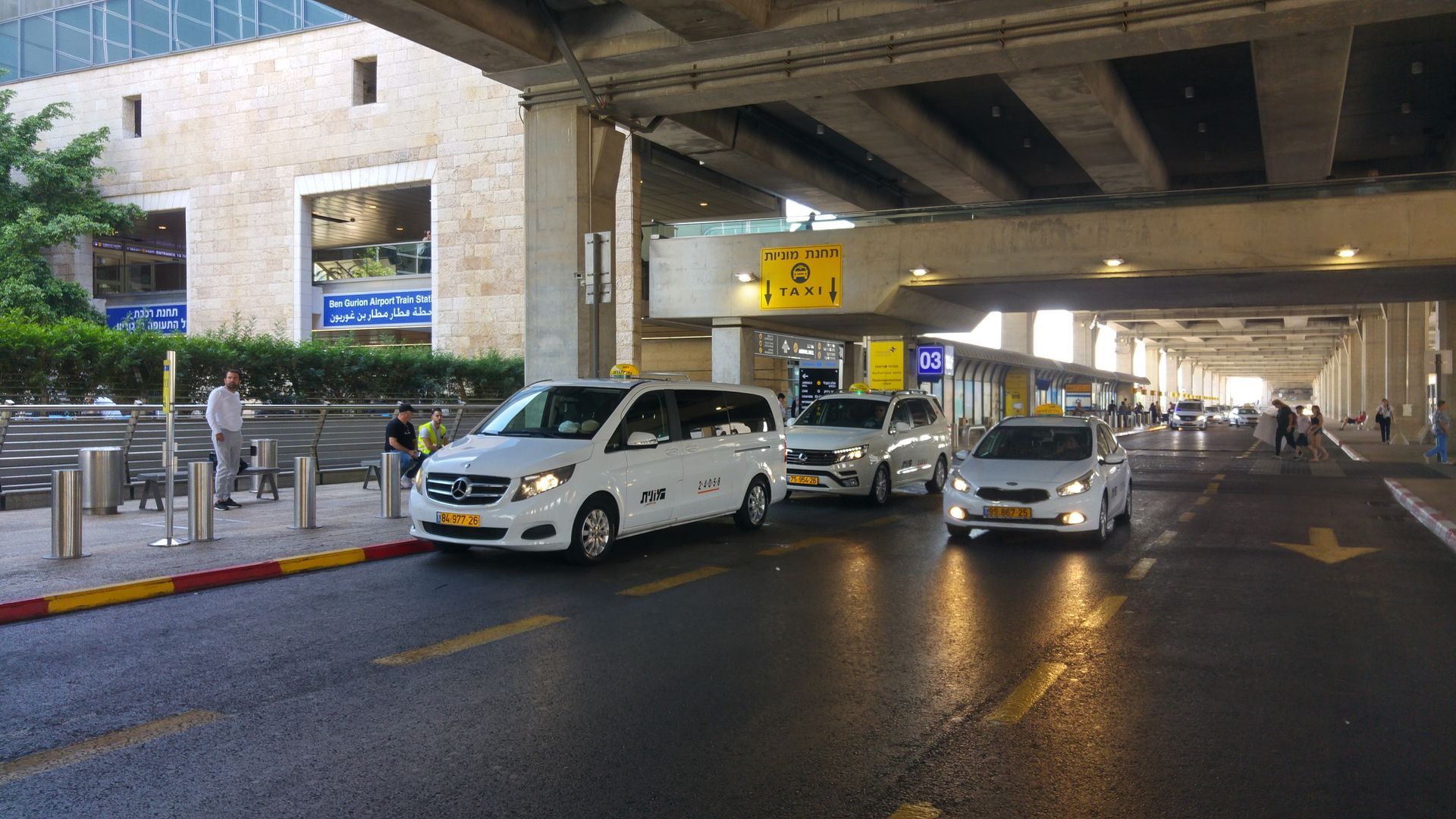 A row of white cars are driving down a street under a bridge.