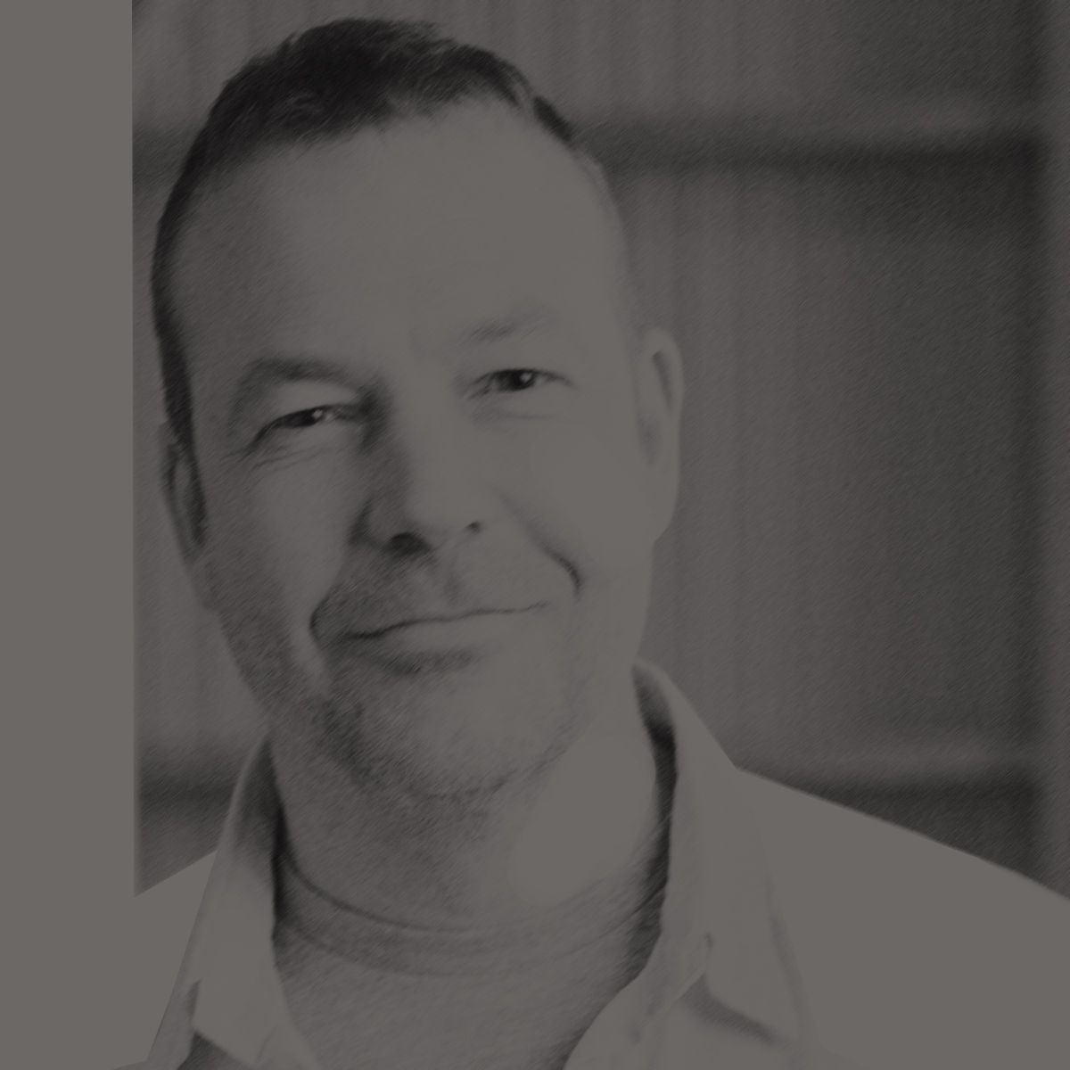 Un homme en chemise claire sourit dans un bâtiment aux poutres apparentes.