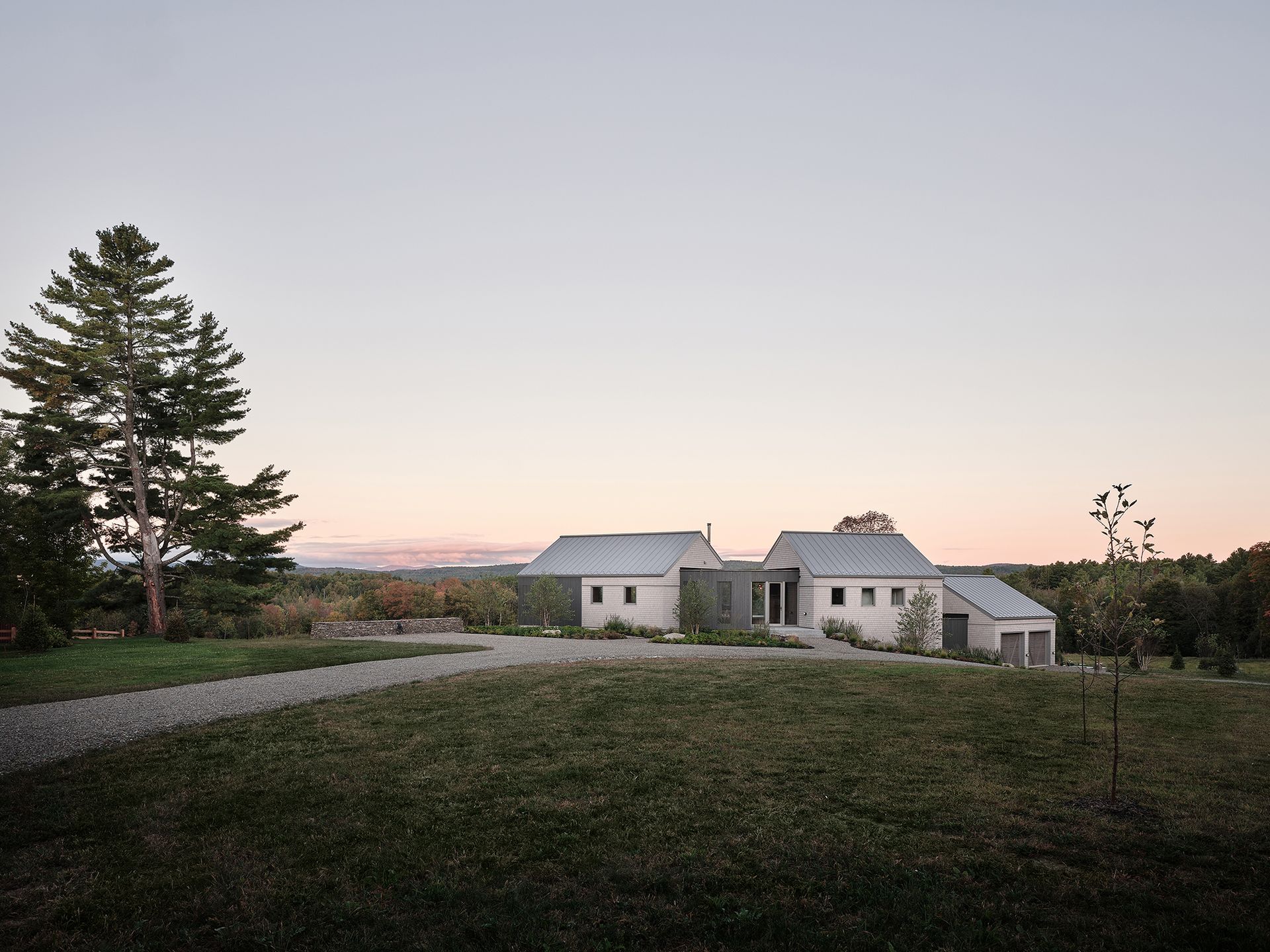 Une maison en pierre claire avec une allée de gravier, située dans un champ herbeux sous un ciel crépusculaire.