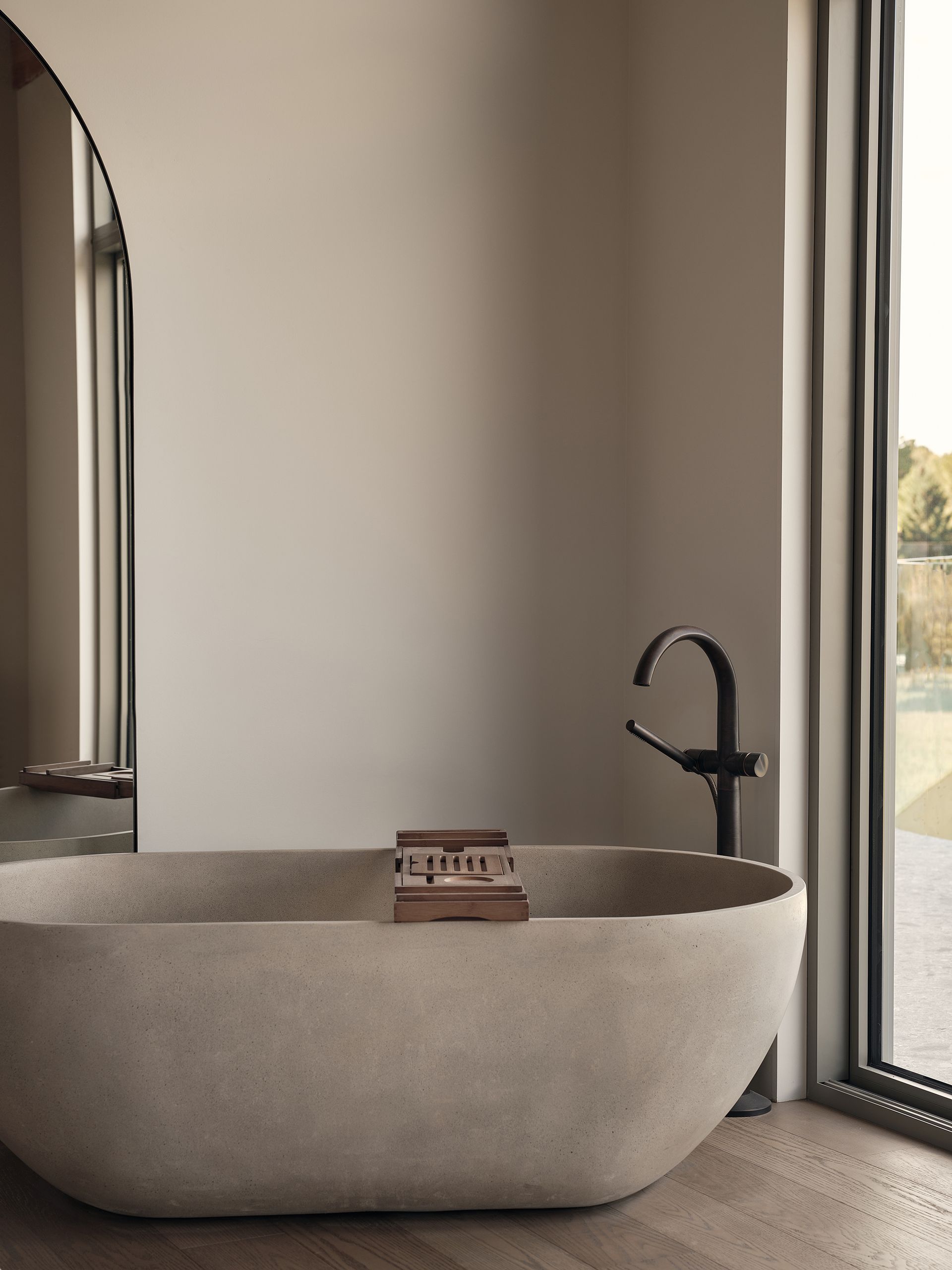 Baignoire îlot beige avec plateau en bois, robinet noir et miroir arqué dans une salle de bains aux tons neutres.