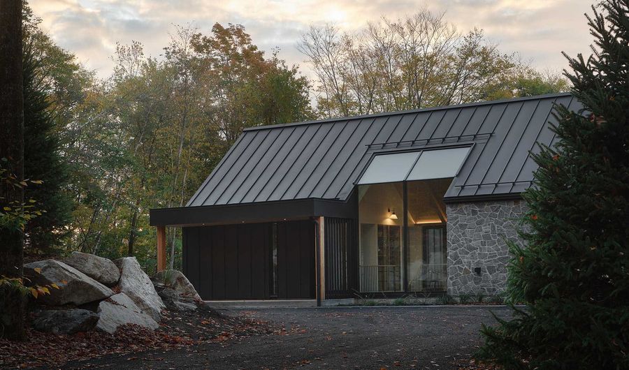 Maison moderne en noir et blanc avec une femme passant devant un abri de voiture par une journée ensoleillée.