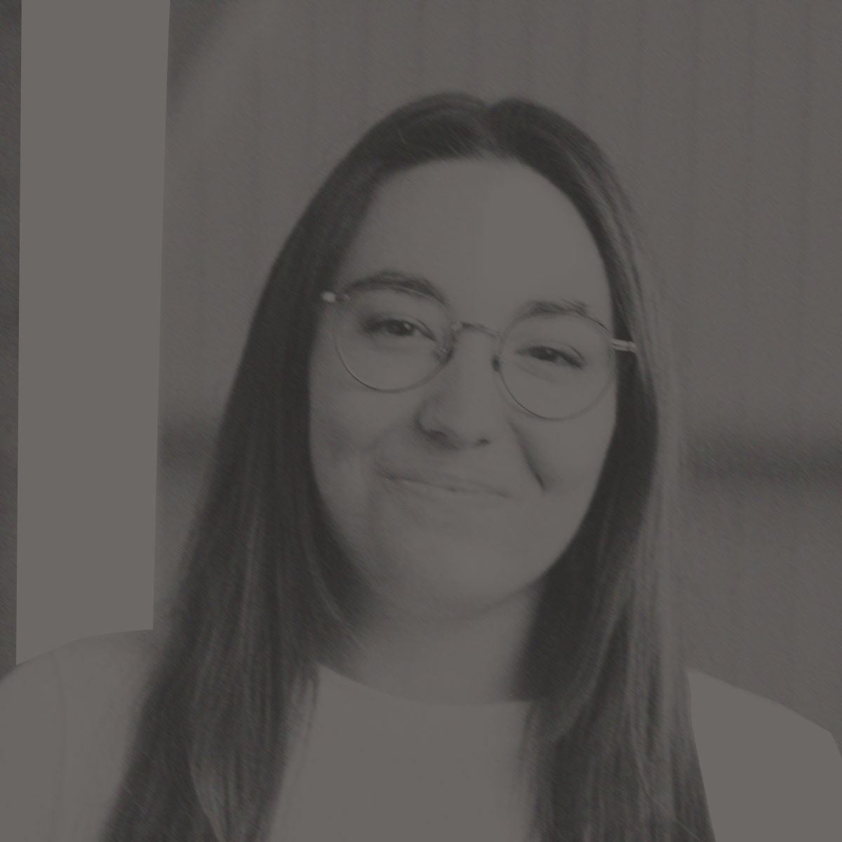 Femme souriante portant des lunettes et un t-shirt blanc. Longs cheveux bruns, à l'intérieur, poutres apparentes.