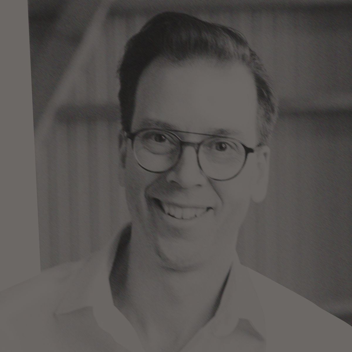 Un homme à lunettes sourit, vêtu d'une chemise blanche ; la scène se déroule dans un bâtiment en bois et en tôle ondulée.