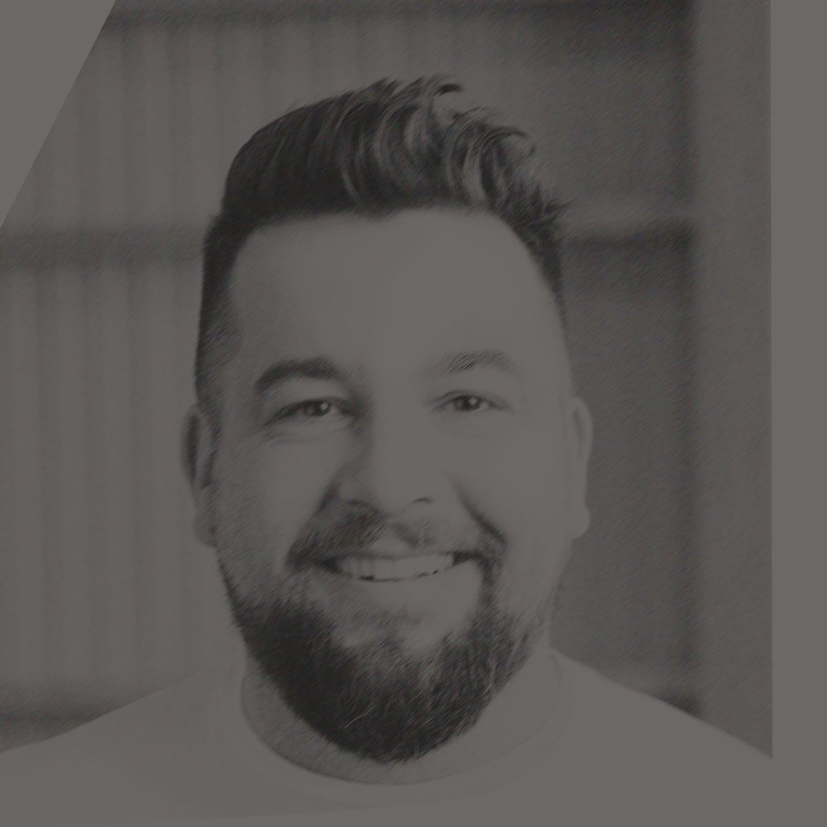 Un homme aux cheveux et à la barbe foncés sourit, vêtu d'une chemise bleu clair ; dans un bâtiment rustique.