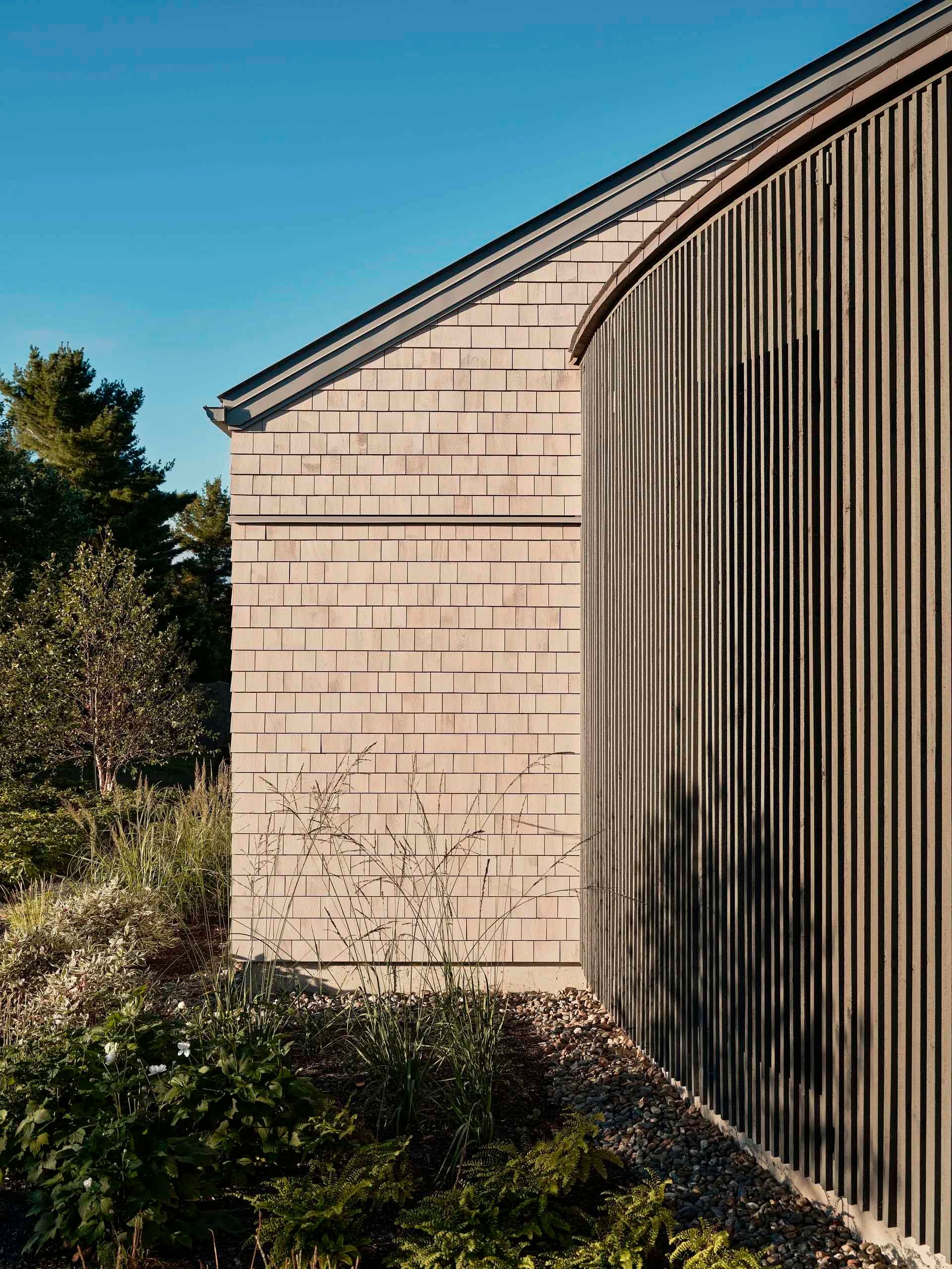 Extérieur d'un bâtiment avec un bardage en bois patiné et un mur arrondi à panneaux verticaux, à côté de verdure sous un ciel bleu.