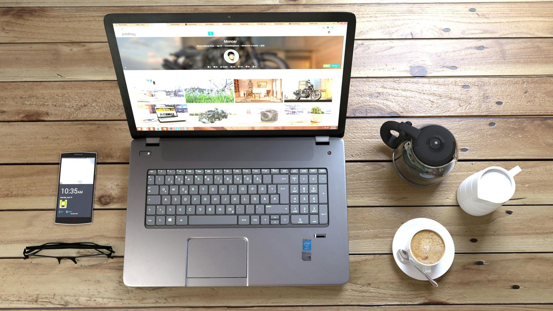 Ein grauer Laptop auf einem rustikalen Holzschreibtisch, daneben ein kleines Gefäß, Gläser, eine Kaffeekanne und eine Tasse Kaffee.