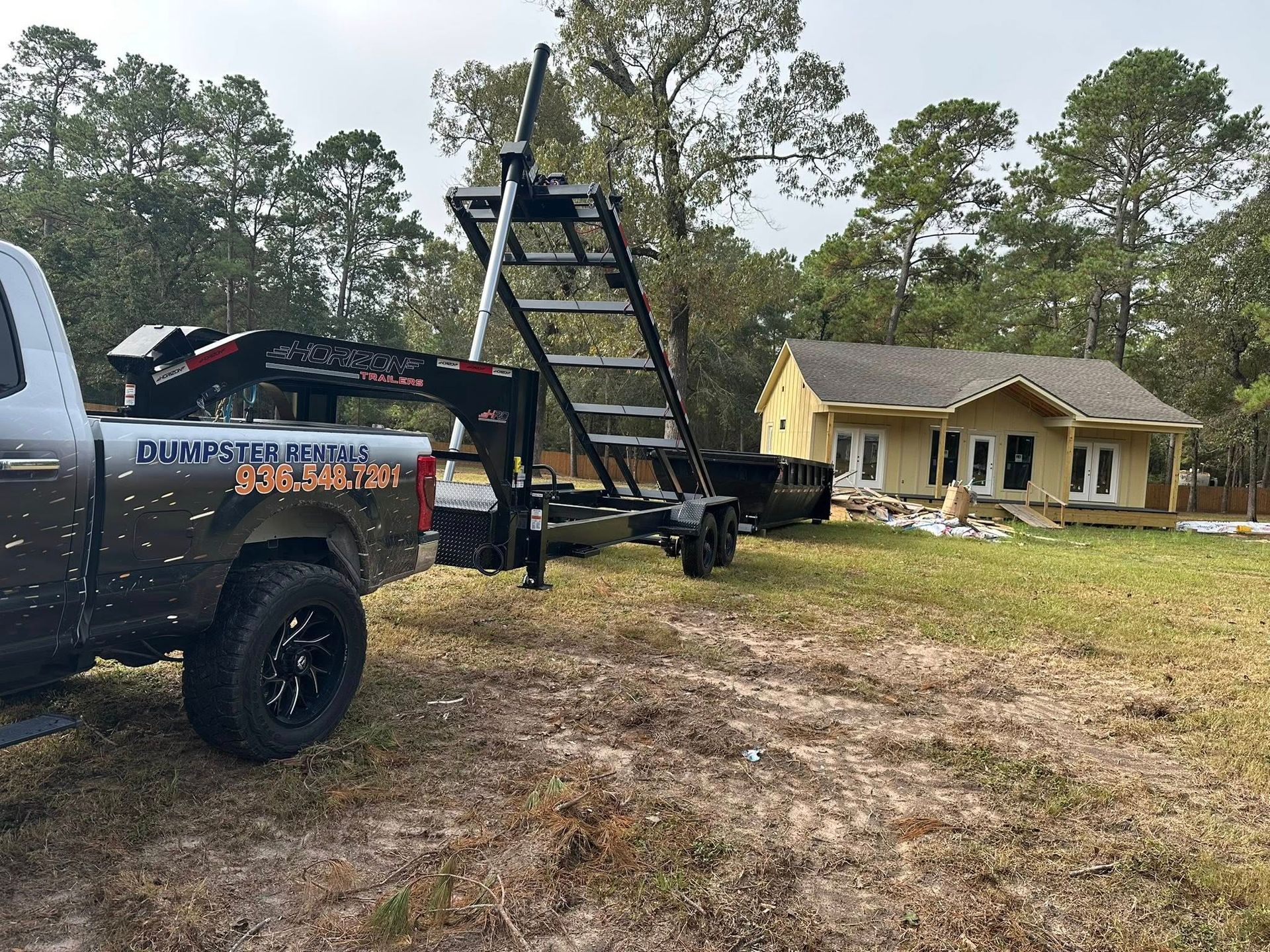 Truck towing a trailer with a tall ladder, near a partially built house in a grassy yard.