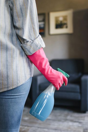 Person wearing pink gloves and holding a blue spray bottle in a room with a sofa.