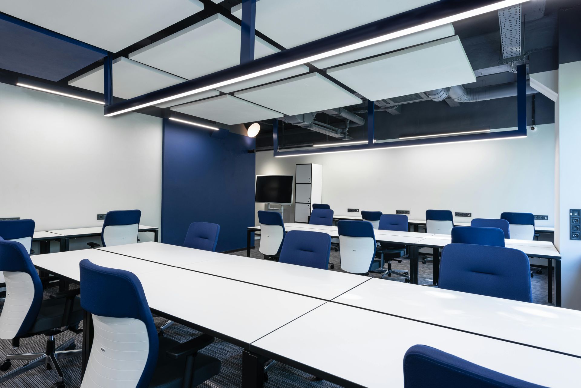 Blue and white classroom with long tables and chairs, under ceiling lights.