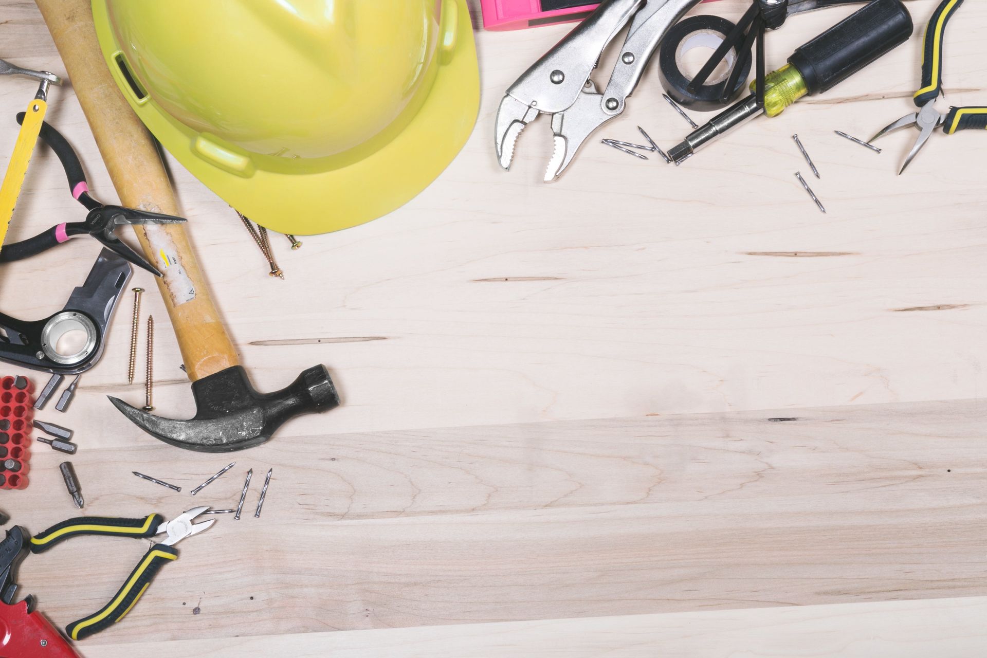Yellow hard hat and various tools (hammer, pliers, tape, nails) arranged on a wooden surface.