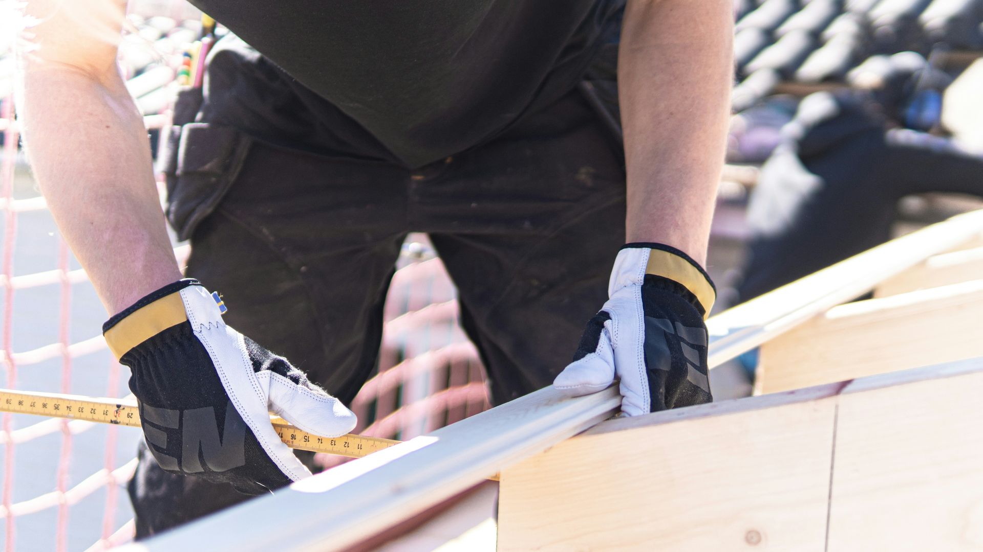 Construction worker measuring wood on a roof, wearing work gloves, another worker in the background.