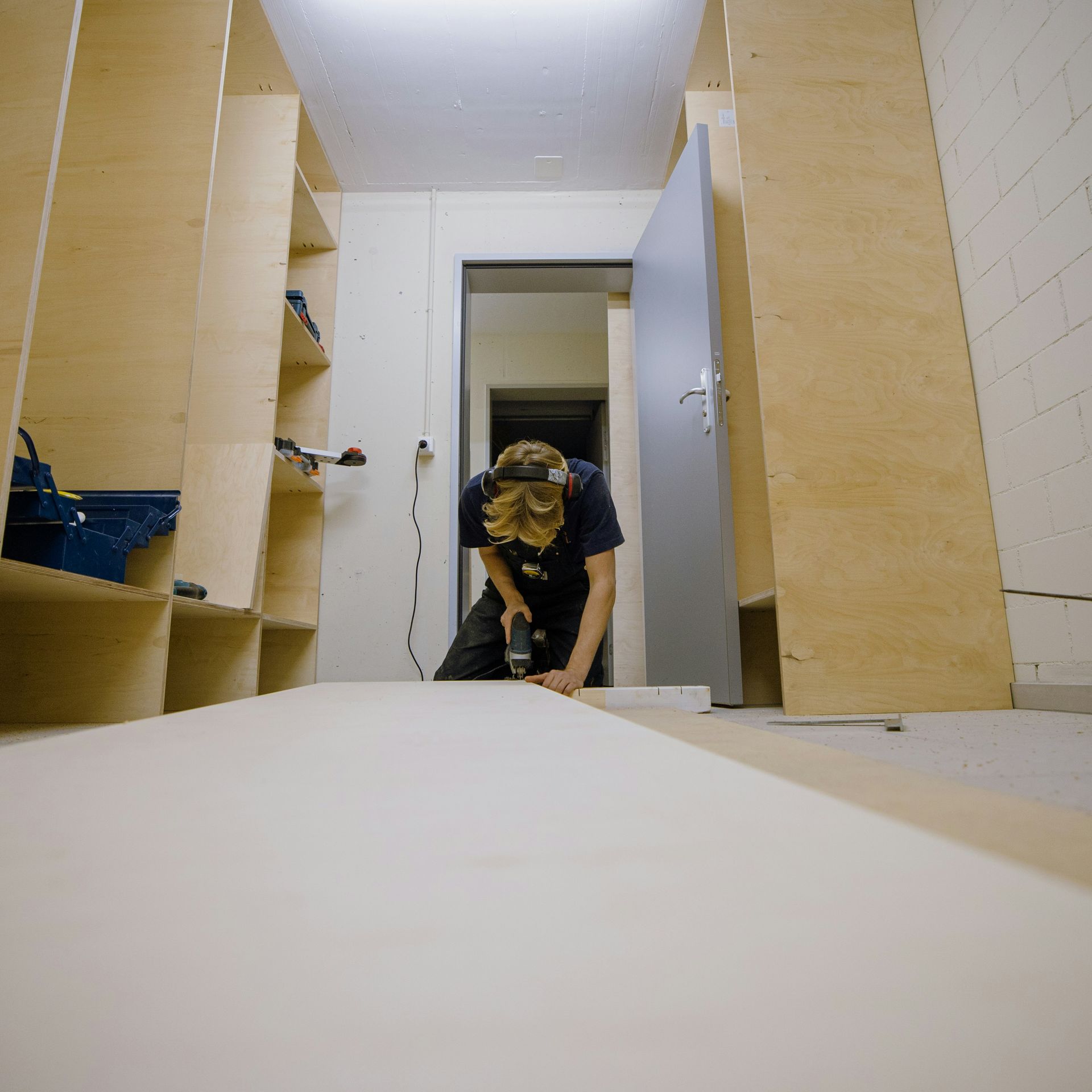 Person using a drill to work on wooden shelves in a room, door open in the background.