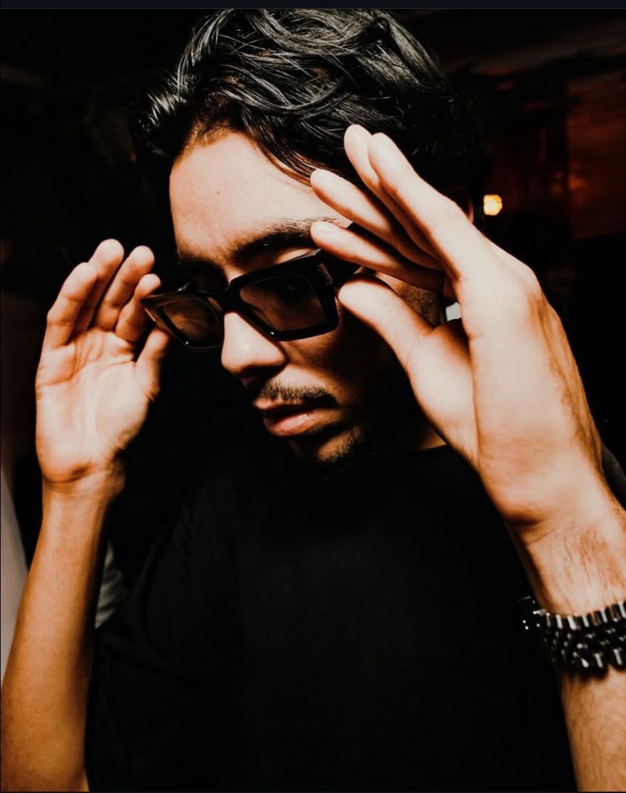 Man in black shirt and sunglasses adjusts them, looking down. Dark setting.