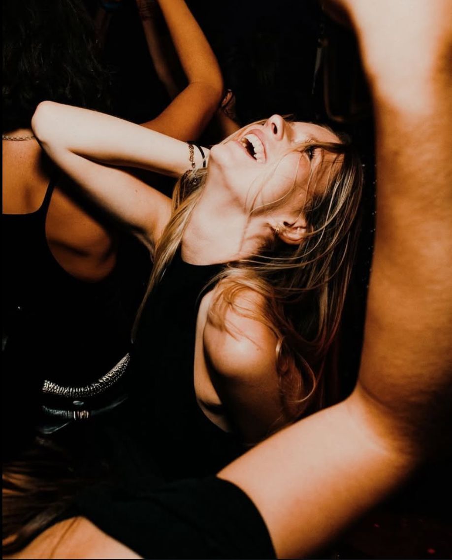 Woman laughing, arms raised, dancing in a dark club. Other people are blurred in the background.