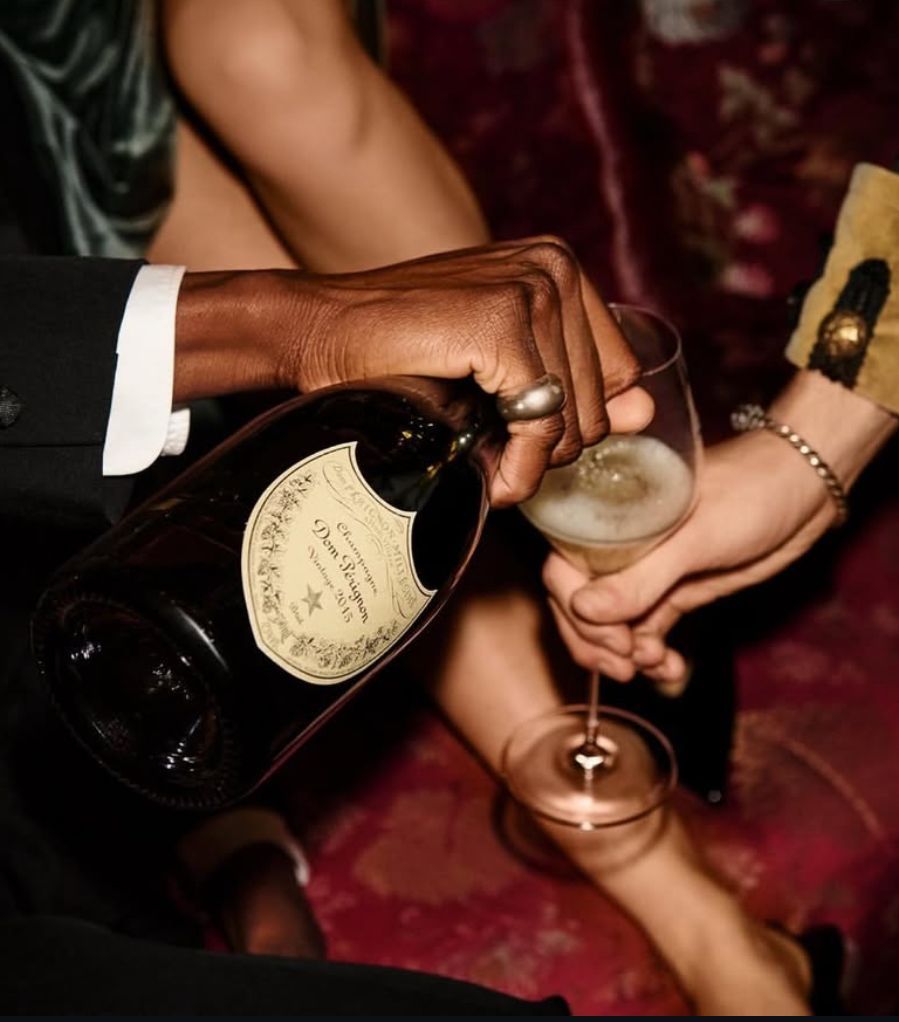 Person pouring champagne into a glass. Dark-skinned hand, Dom Pérignon bottle. Red setting.