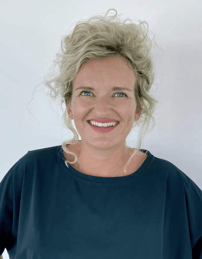 Een vrouw met blond krullend haar draagt ​​een blauw shirt en glimlacht.