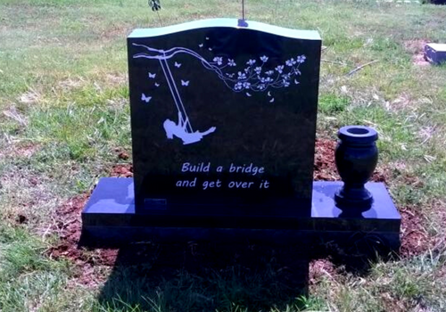 Black memorial headstone with a bridge and  figure on a swing engraving in a grassy cemetery.