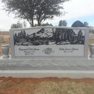 Granite memorial plaque with engraved landscape scene and text, set outdoors on a stone base
