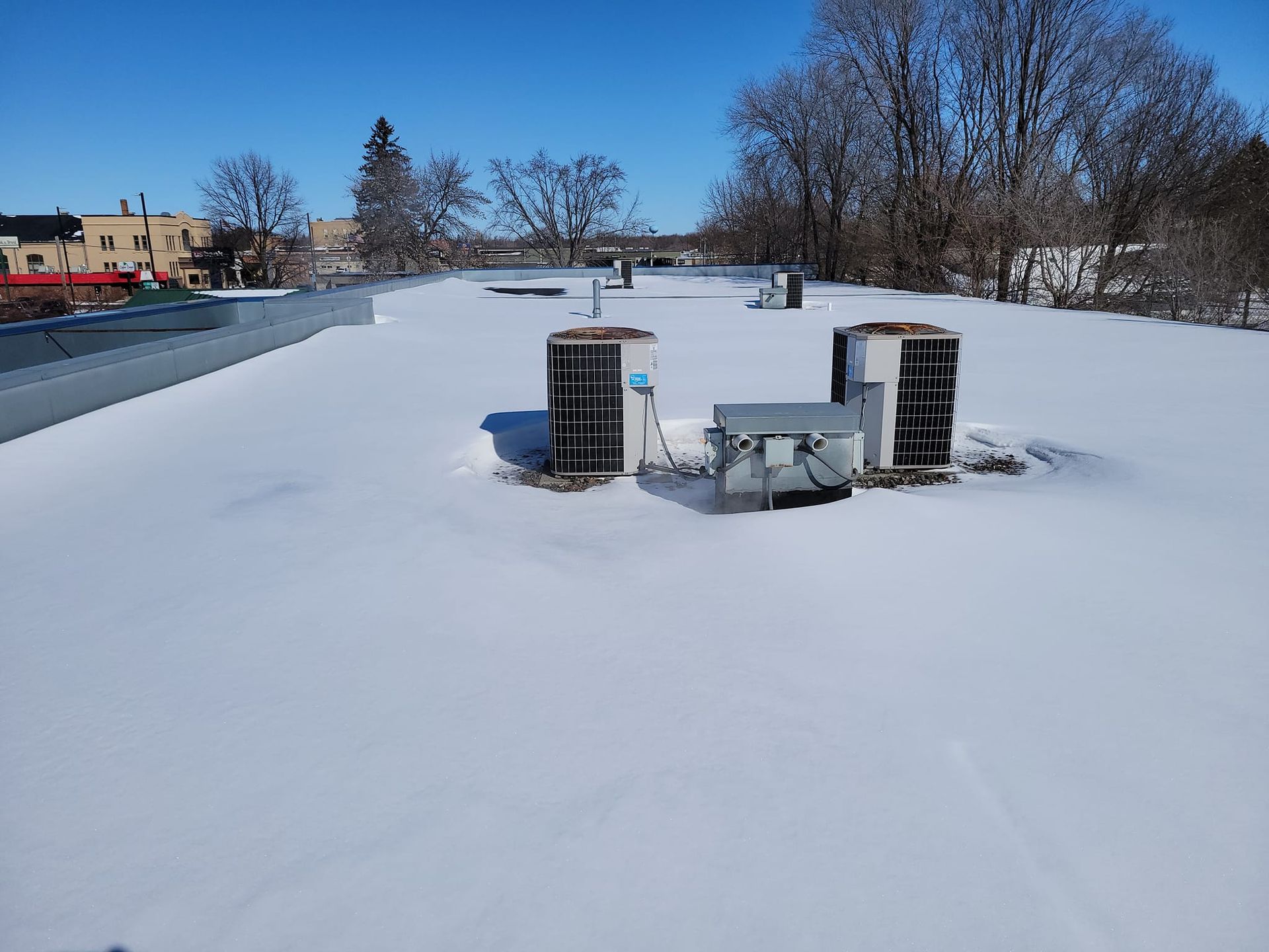 Snow-covered flat roof with vents and equipment, against a backdrop of trees and buildings on a sunny day.