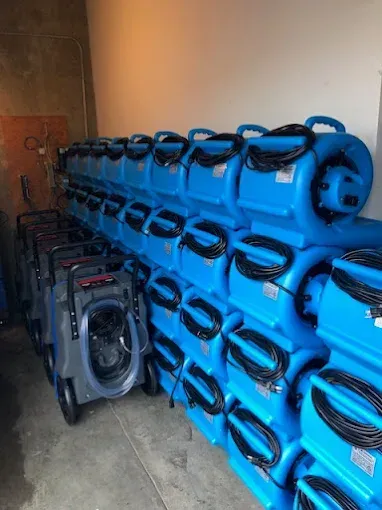 Row of blue water damage fans stacked with a few gray vacuums in a storage room.