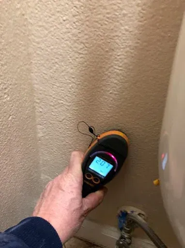 Hand holding a moisture meter against a beige wall, near a white water heater. Display reads 28.7.