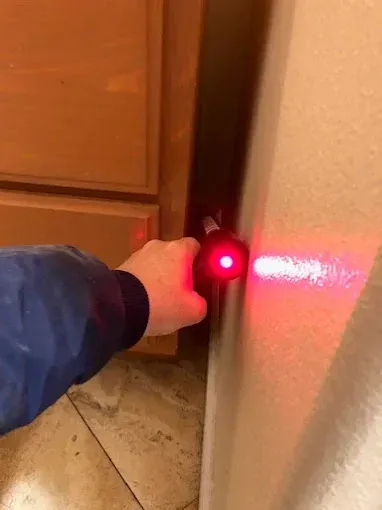 Hand holding a red laser pointer, aiming it at a wall between a cabinet and a wall in a room.