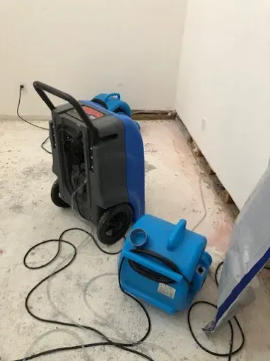 Two blue dehumidifiers in a room with exposed flooring. A cord and handle on the larger one.