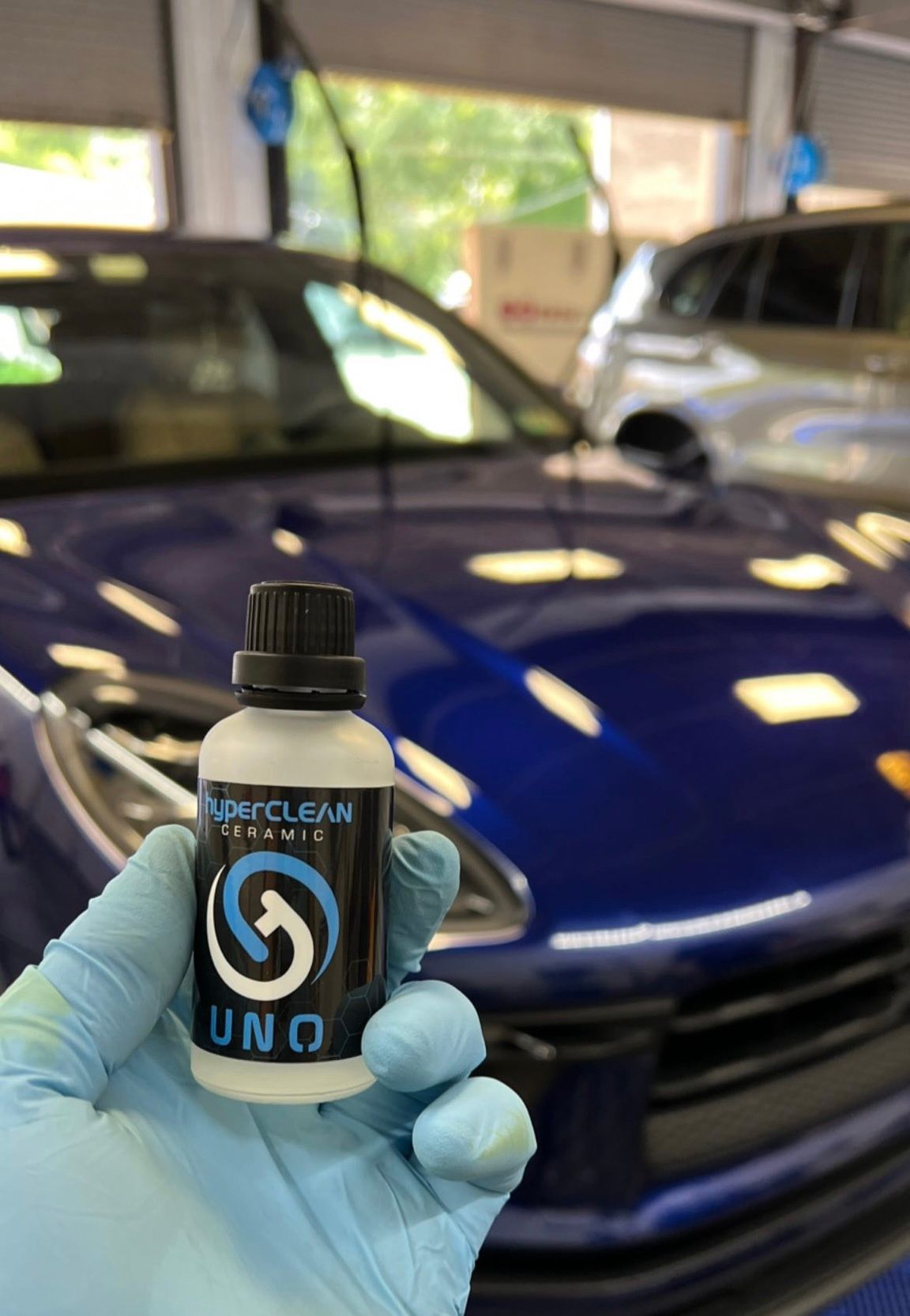 A person is holding a bottle of ceramic coating in front of a blue car.