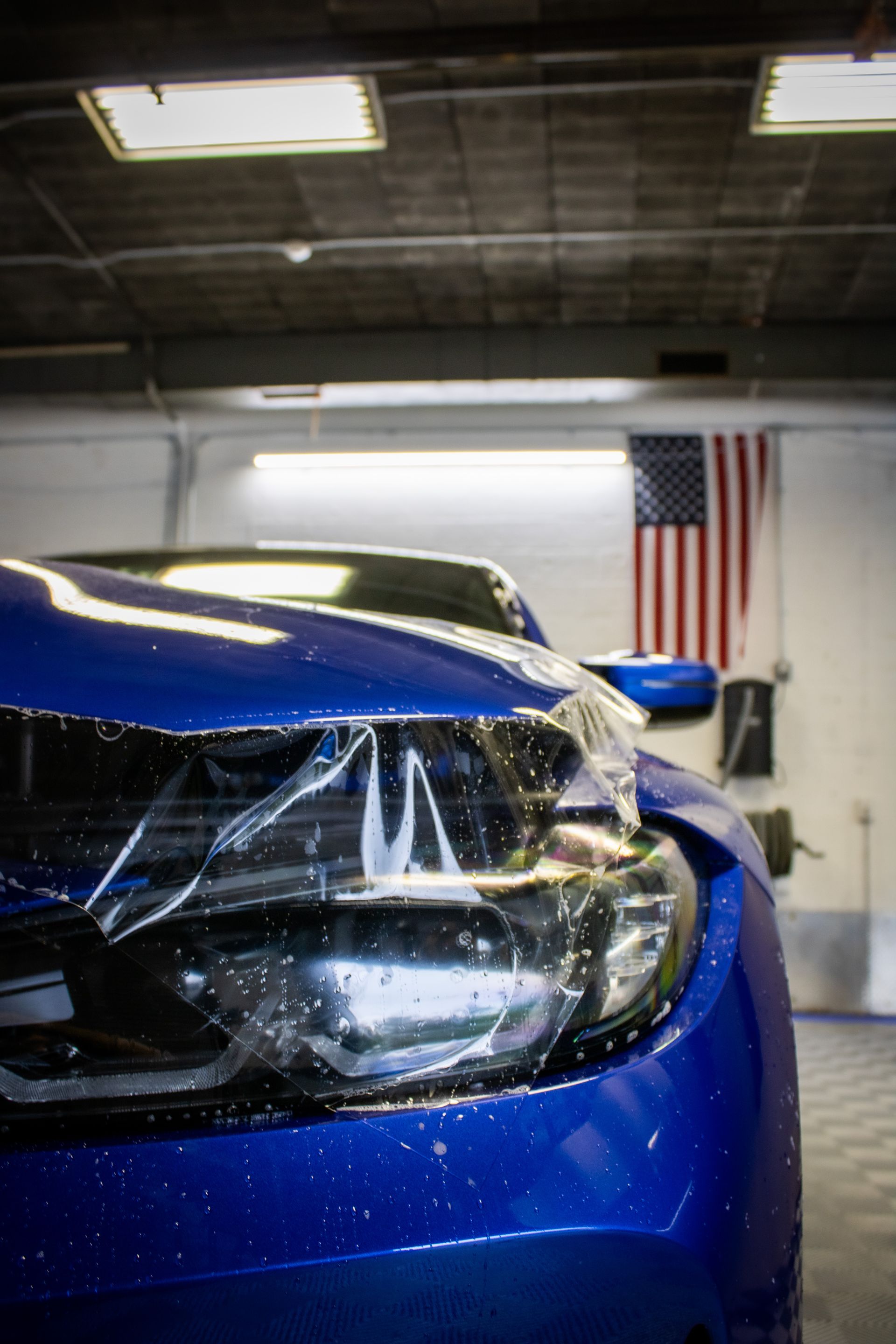Blue sports car front end in a garage, with an American flag in the background.