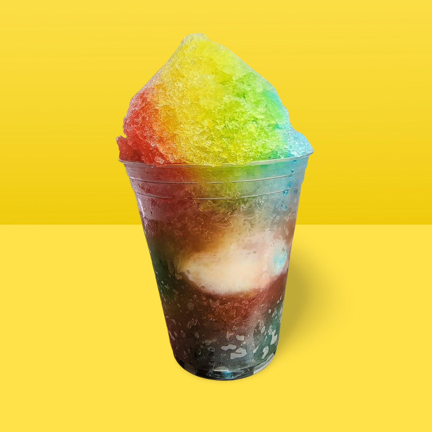 A rainbow colored shaved ice drink in a plastic cup on a yellow table.