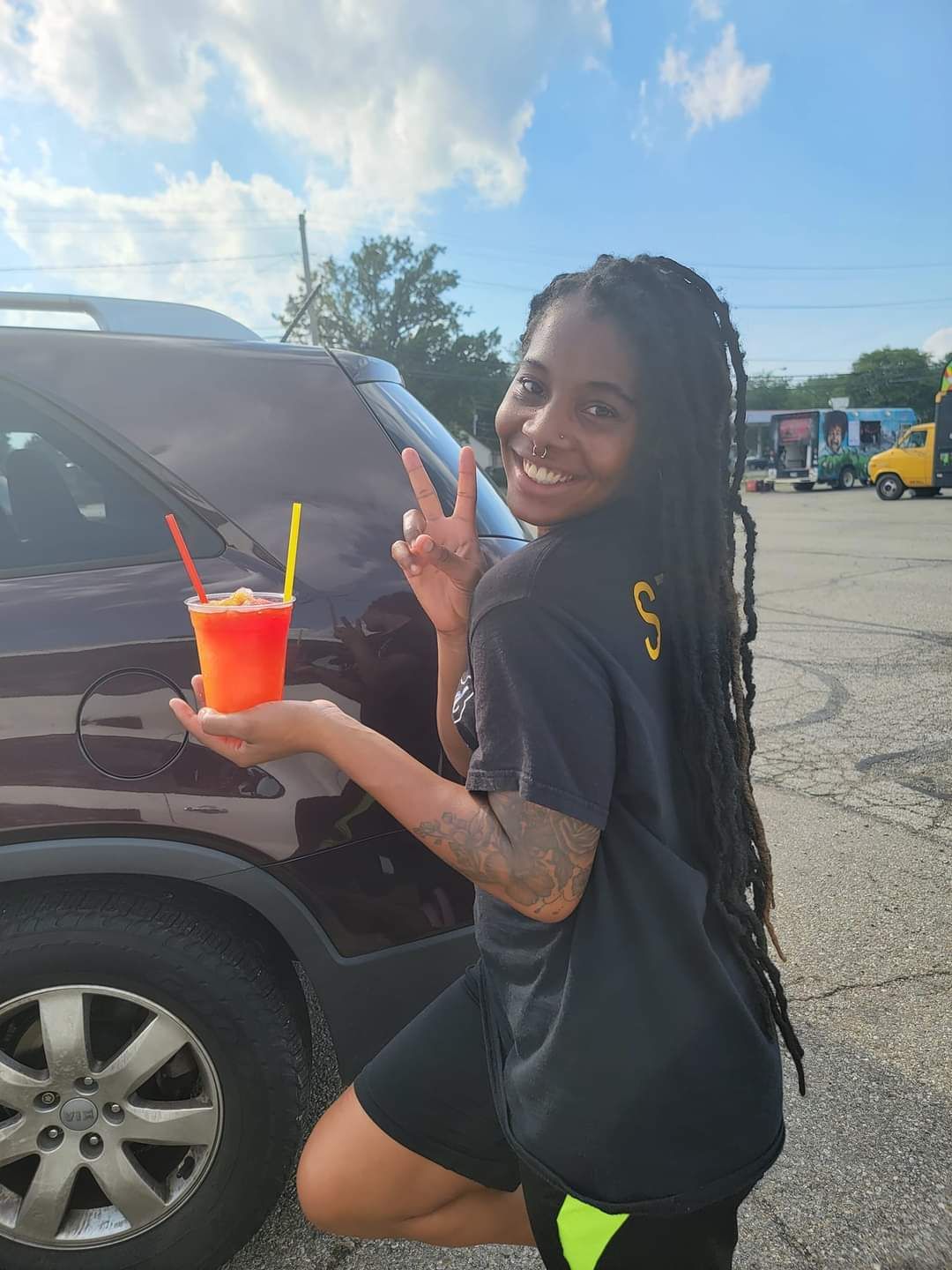 A woman is kneeling down in front of a car holding a drink and giving a peace sign.