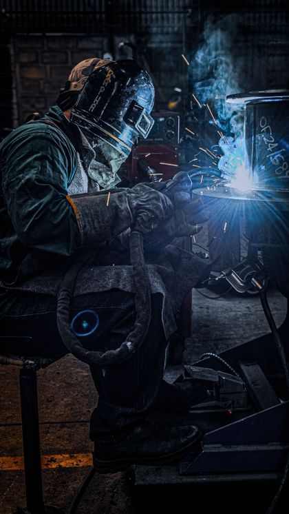 Un soldador con equipo de protección produce una chispa de luz azul mientras trabaja con metal en un taller oscuro.
