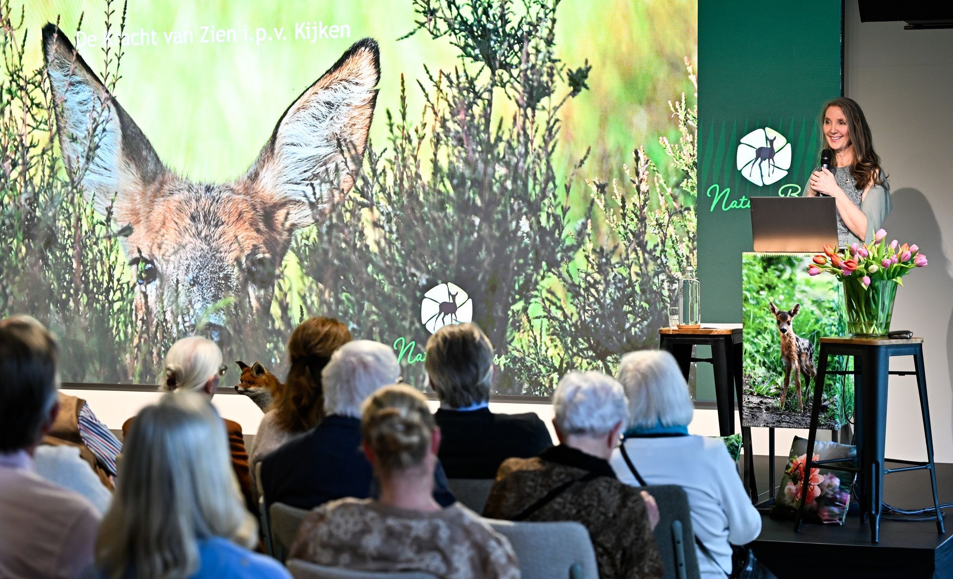 Vrouw geeft een presentatie over paddenstoelen aan een publiek; geprojecteerd beeld van paddenstoelen.
