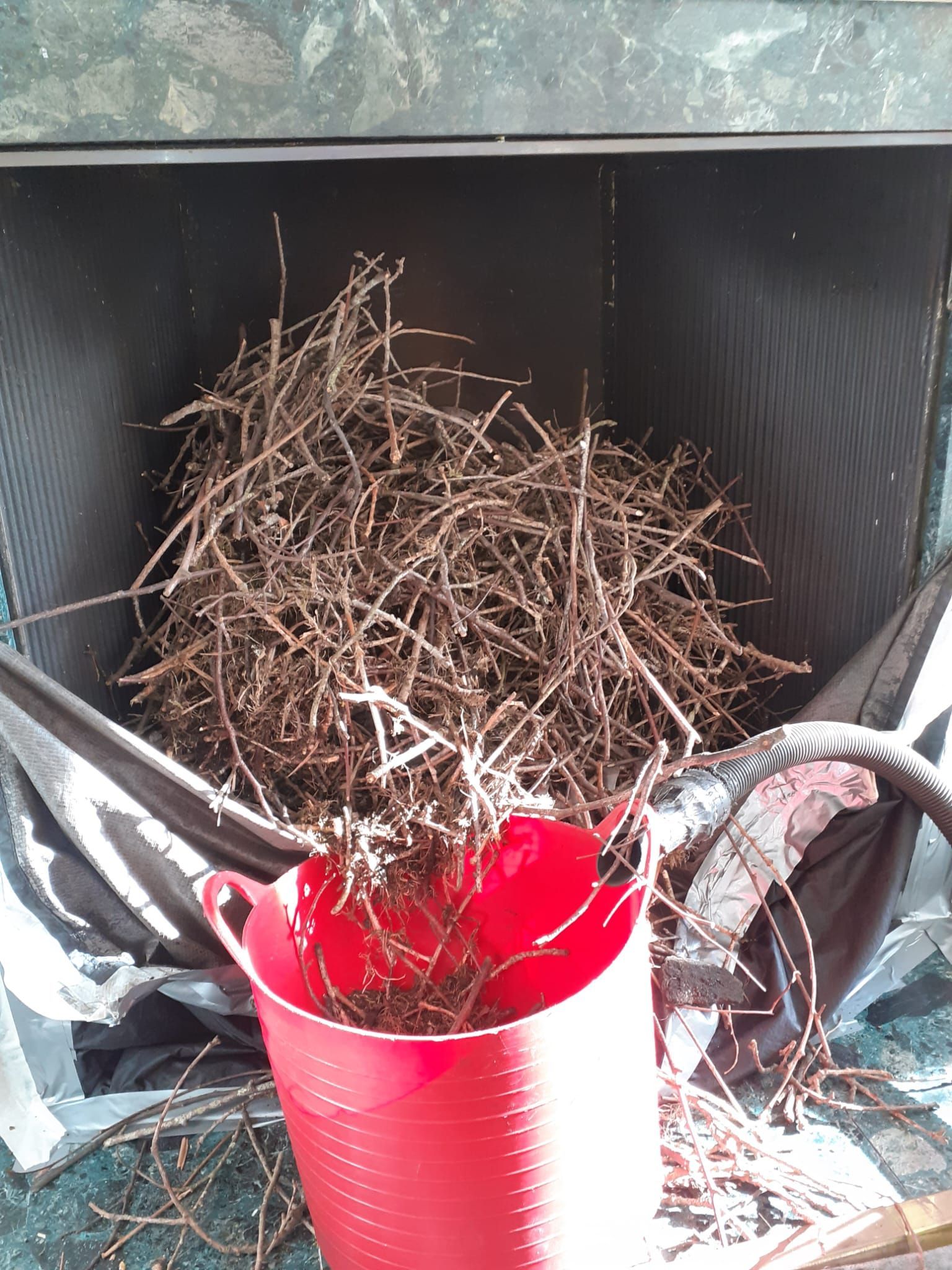 A red bucket filled with small brown branches sits in front of a black container filled with more branches.
