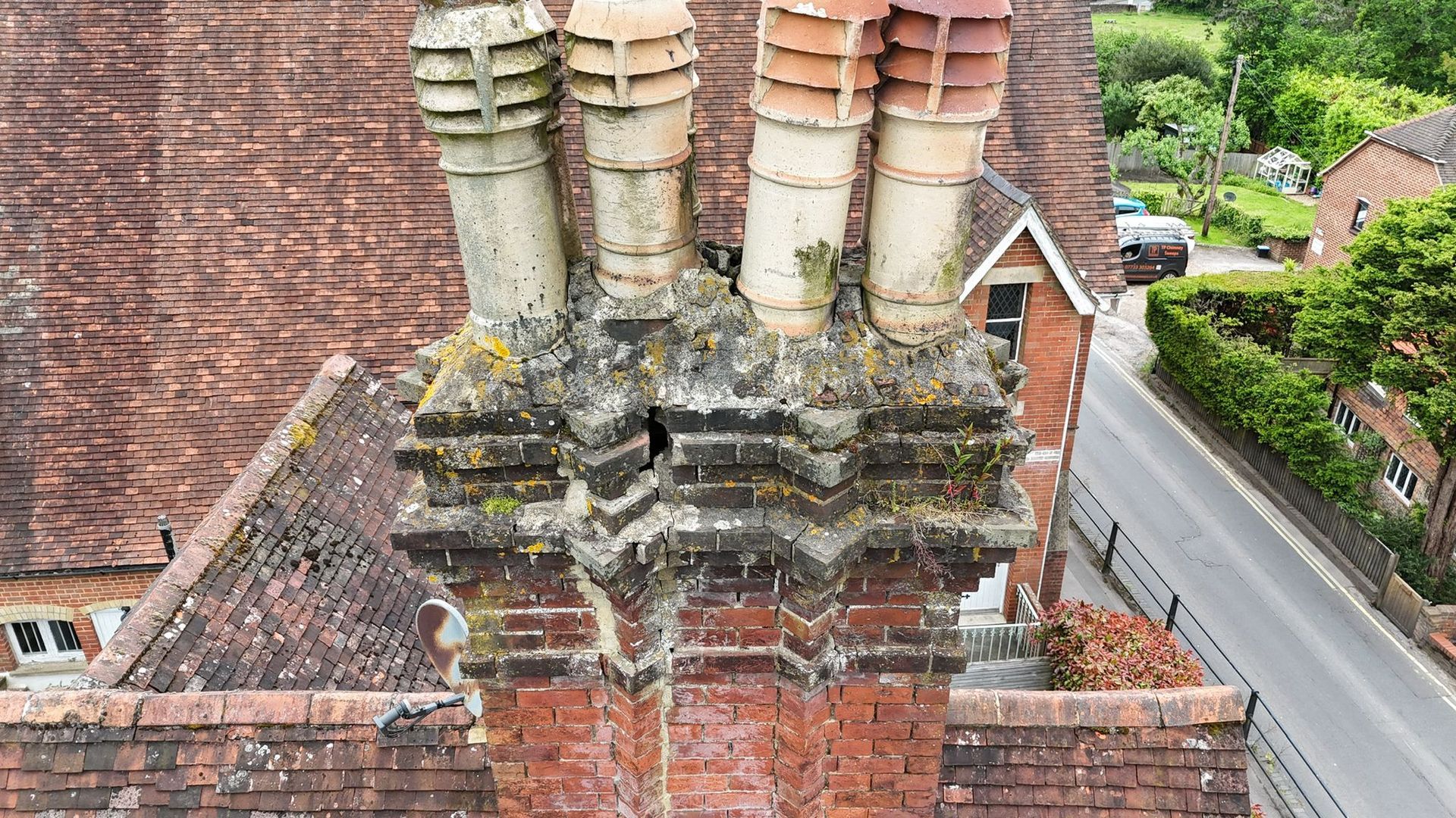 Brick chimney with four fluted chimney pots on a tiled roof, overlooking a street and green foliage.