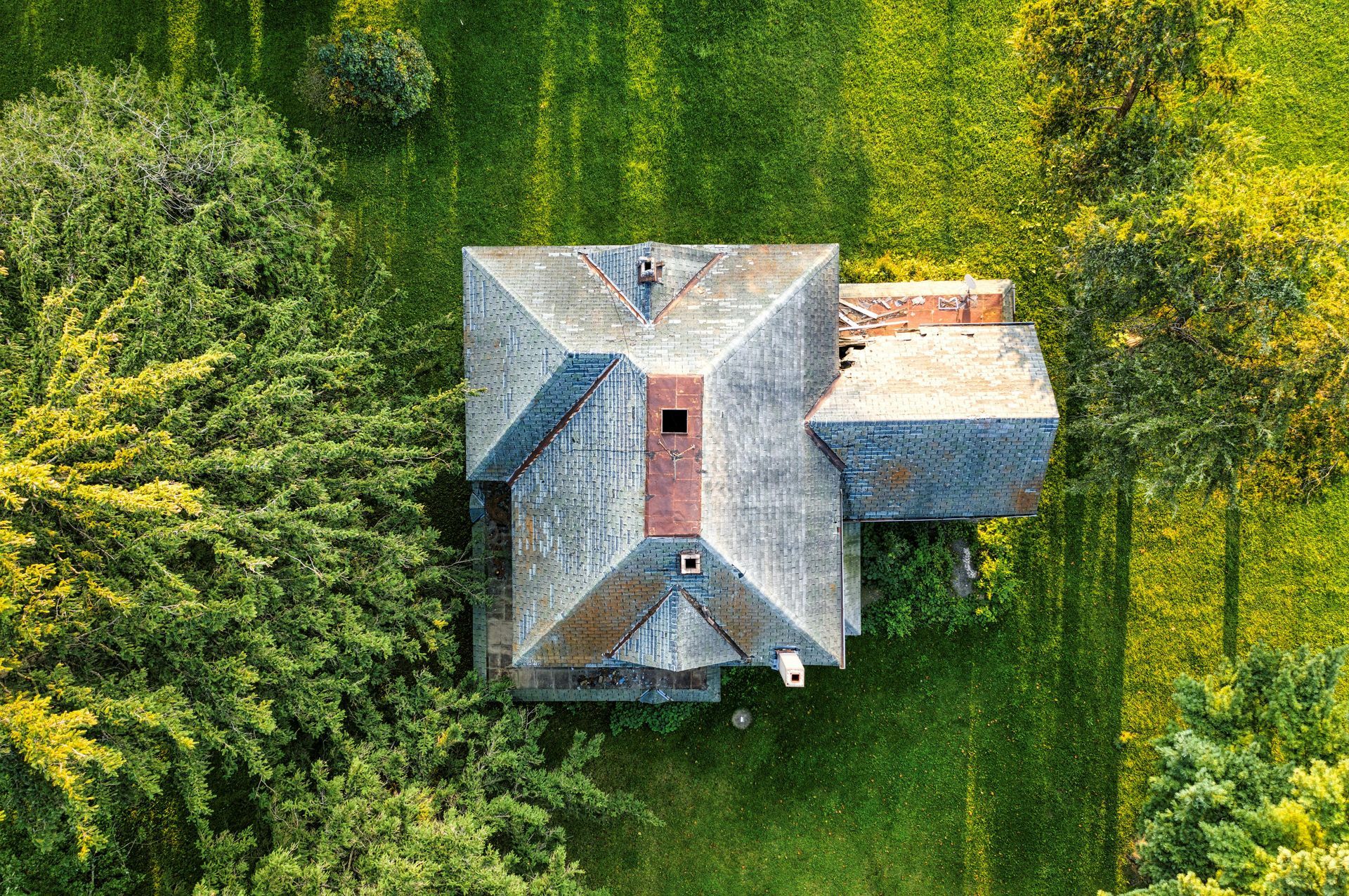 Overhead view of a house with a weathered gray roof, surrounded by green trees and grass.