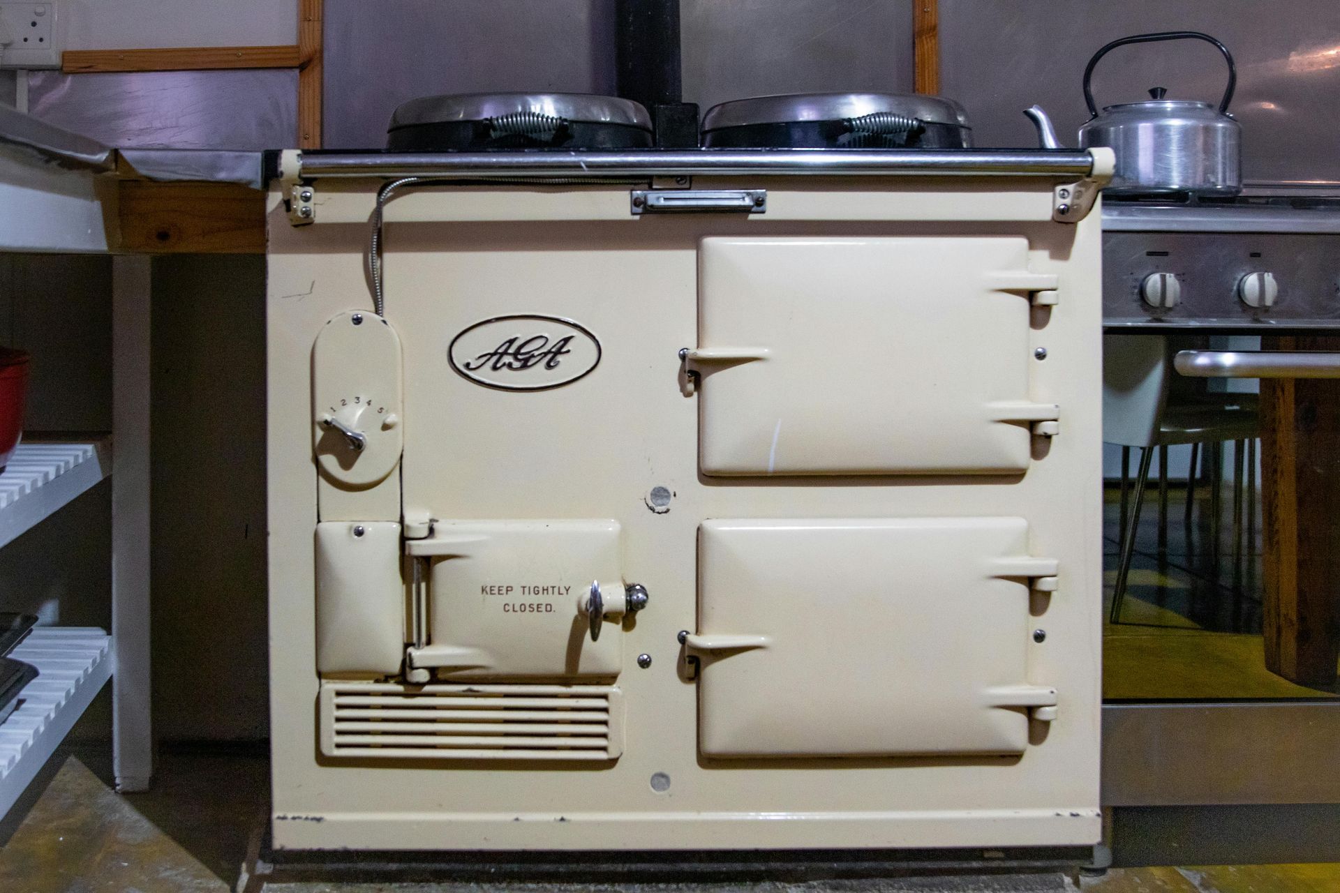 Cream-colored Aga range cooker with black top burners in a kitchen setting, next to a stainless steel gas stove.