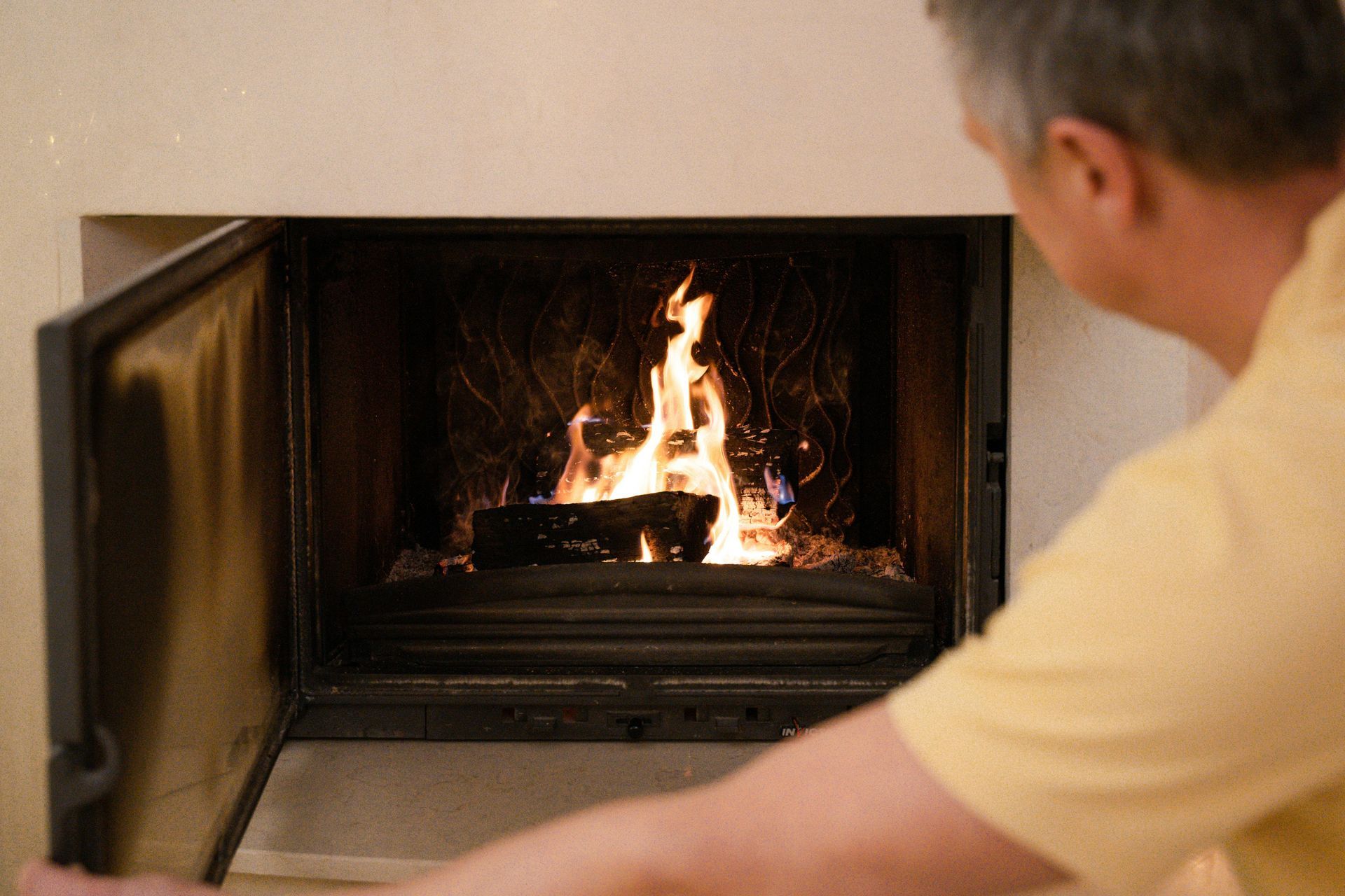 Man opening a fireplace door with burning logs inside. The man wears a yellow shirt in a neutral-toned room.