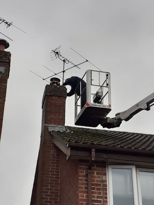 A person in a lift bucket adjusts a TV antenna atop a brick chimney on a cloudy day.