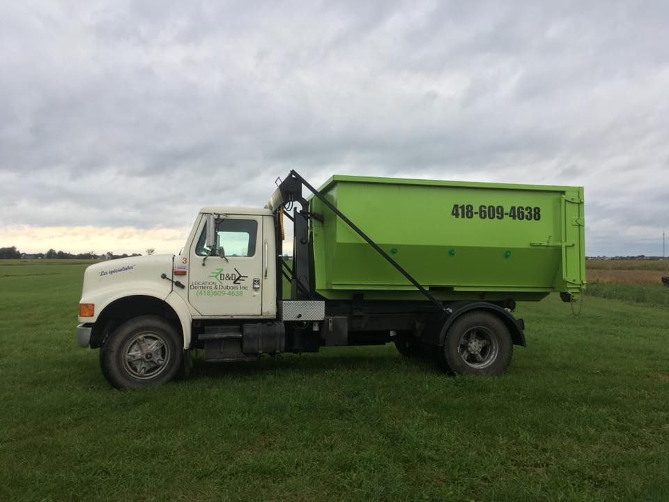 Camion blanc avec benne verte dans un champ herbeux. Numéro de téléphone sur le côté de la benne.
